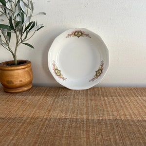 May include: White ceramic bowl with a floral design in pink, yellow, and green. The bowl has a scalloped edge and is sitting on a wooden surface.