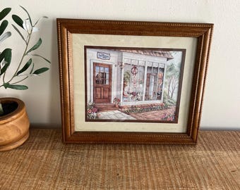 May include: A framed print of a quaint shop with a brown door and a sign that reads "The Flower Shop". The shop has a large window with a view of flowers and other items inside. The frame is brown wood with a decorative border.