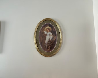 May include: A framed oval print of a young girl in a white dress and straw hat holding a bouquet of flowers. The frame is gold with a decorative design.