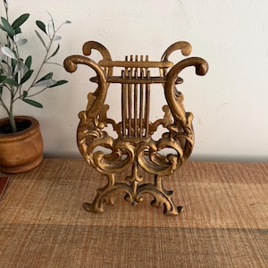 May include: A gold-colored metal lyre-shaped object featuring ornate, swirling scrollwork details and vertical bars across the center. The structure has a wide, decorative base and curved arms extending upward, finished with a textured, metallic gold patina.