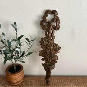May include: A bronze-coloured wall sconce with a floral and ribbon design. The sconce features a detailed arrangement of flowers and a decorative bow at the top. It includes a small cup for holding a candle. A small olive tree in a wooden pot is to the left.