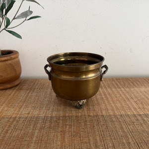 May include: A small, round, polished brass pot featuring two side handles and three distinct feet. The metal surface has a warm, golden-brown tone with a smooth finish. The vessel has a wide, open top and a curved body, showcasing a classic metallic construction.