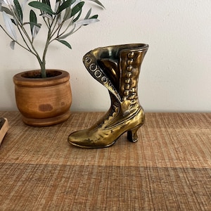 May include: A vintage brass boot-shaped vase. The boot has a high heel and is detailed with buttons and decorative stitching. The vase is approximately 6 inches long and 5 inches tall. It is a decorative item, likely for small flowers or display.