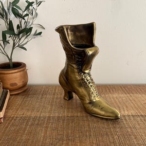 May include: A vintage-style, gold-colored boot-shaped vase. The boot features a pointed toe, a high heel, and a lace-up design. The vase is made of metal and sits on a wooden surface. A small potted plant and books are in the background.