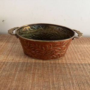 Op de afbeelding: Een ovale koperen kom met een reliëfpatroon van bladeren op zowel de buiten- als de binnenkant. De kom is voorzien van twee decoratieve handvatten aan weerszijden, met een warme, metalen afwerking en zichtbare sporen van een verouderde patina.