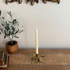 Puede incluir: Un candelero de hoja de oro con una sola vela blanca. El candelero está sobre una superficie de madera con una pila de libros.