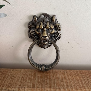 Op de afbeelding: Een metalen deurklopper in bronskleur met een gedetailleerde leeuwenkopsculptuur en vloeiende manen. Een grote, cirkelvormige ring hangt aan de bek van de leeuw, ontworpen om tegen een oppervlak te slaan wanneer deze wordt opgetild en losgelaten.