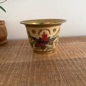 May include: A small, brass-colored decorative bowl with a wide rim. The bowl features a cream-colored background with a floral design in red, green, and brown. The design includes a candle and pinecones. The bowl is sitting on a wooden surface.