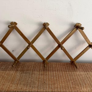 May include: A wooden expanding coat rack with multiple pegs, made of light brown wood. The rack is mounted on a white wall. The design allows the rack to expand and contract, providing adjustable storage space for coats, hats, or other items.
