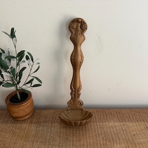May include: Ornate, bronze-colored serving spoon with a decorative handle and a shallow, rounded bowl. The spoon features intricate carvings and a vintage aesthetic, suitable for serving food. The spoon is displayed on a wooden surface.