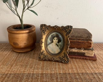 May include: A gold-toned ornate frame with a portrait of a woman with dark hair and a white dress. The frame is sitting on a stack of old books.