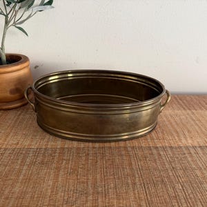 May include: An antique brass oval planter with two small handles. The planter is empty and sits on a wooden surface. A small olive tree in a wooden pot is in the background.