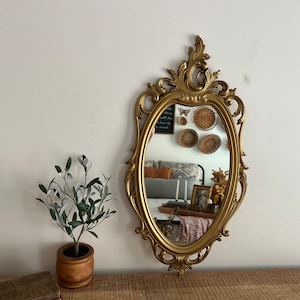 May include: A gold ornate oval mirror with a decorative frame. The mirror reflects a room with a sofa, cushions, and a table with candles and a vase.
