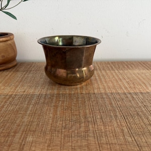 Puede incluir: Un pequeño cuenco de metal color latón con base redondeada y borde acampanado y festoneado. El cuenco presenta una superficie pulida y reflectante con sutiles variaciones de tono, mostrando una silueta curva y lisa con una boca ancha.