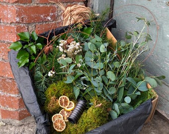 Op de afbeelding: Een doos gevuld met groen, waaronder eucalyptus, dennen en mos. Gedroogde citroenschijfjes en een draadspoel zijn tussen het gebladerte genesteld. De doos is bekleed met zwart papier, wat een natuurlijke opstelling creëert.