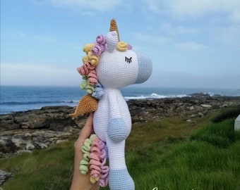 Puede incluir: Un juguete de unicornio de crochet con cuerpo blanco, patas azules y una melena y cola arcoíris. El unicornio está siendo sostenido en una mano contra un fondo de costa rocosa y océano.