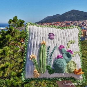 Puede incluir: Una almohada blanca de crochet con un diseño de cactus verde, amarillo y morado. La almohada tiene un borde de crochet verde y blanco.