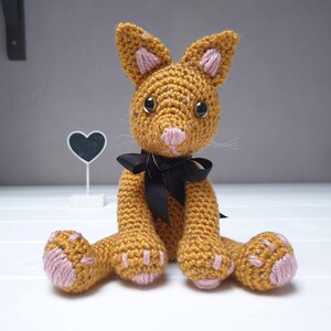 May include: A crocheted stuffed animal cat with a brown and orange body, pink ears and paws, and black eyes. The cat is wearing a black ribbon around its neck.