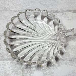 May include: Clear glass leaf-shaped dish with a scalloped edge and a small handle. The dish has a ribbed design radiating from the center, creating a textured appearance. The clear glass allows light to pass through, highlighting the intricate details.