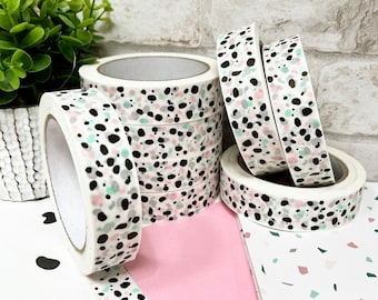 May include: A stack of white washi tape rolls with a black, pink, and green speckled pattern. The tape is on a white surface with a pink and white patterned paper and a white paper with a gray, pink, and green geometric pattern.