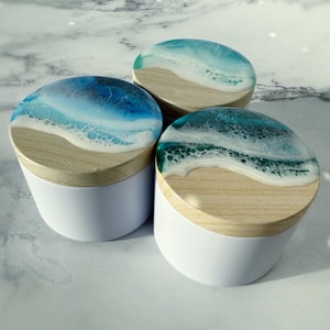 Könnte beinhalten: Drei weiße und holzfarbene Behälter mit Deckeln aus Harz zum Thema Ozean. Die Deckel weisen ein Wellenmuster in Blau-, Weiß- und Türkistönen auf, das einer Miniatur-Meereslandschaft ähnelt. Die Behälter sind auf einer Marmoroberfläche angeordnet.