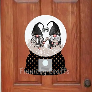 May include: A Halloween-themed snow globe door hanger with two gnomes. The gnomes are dressed in black and white with skeleton and skull details. The snow globe is decorated with black and gold polka dots.