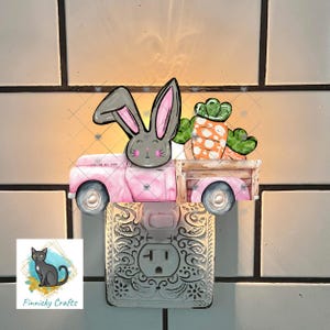May include: A decorative night light featuring a pink truck carrying a gray bunny and carrots. The bunny has pink inner ears and a smiling face. The night light has a white, ornate base with a standard electrical outlet.