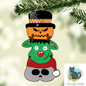 以下が含まれることがあります： ハロウィンをテーマにしたクリスマスオーナメントで、かぼちゃ、緑のモンスター、サンタの帽子をかぶった頭蓋骨が積み重ねられたデザインになっています。かぼちゃは怖い顔をしていて、紫と白のドット模様の黒いシルクハットをかぶっています。モンスターは赤い鼻と顔に縫い目があります。頭蓋骨は灰色で黒い目があります。