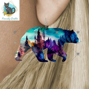 May include: A single bear-shaped earring with a watercolor design of a forest scene in shades of blue, purple, and orange. The earring is hanging from a silver hook. The earring is a product of Finnicky Crafts.