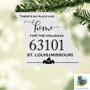 May include: White wooden ornament shaped like the state of Missouri with the text "THERE'S NO PLACE LIKE home FOR THE HOLIDAYS 63101 ST. LOUIS MISSOURI" in black lettering. The ornament has a small string for hanging.