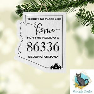 May include: A white wooden ornament in the shape of Arizona, with the text "THERE'S NO PLACE LIKE HOME FOR THE HOLIDAYS 86336 SEDONA | ARIZONA". The ornament is hanging from a twine string and is decorated with a house silhouette.