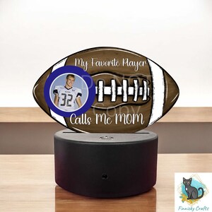 May include: A brown and white football-shaped night light with a photo of a football player in a blue circle. The text "My Favorite Player Calls Me Mom" is written on the football.