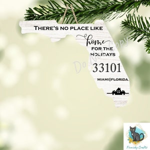 Puede incluir: Adorno de madera blanca con forma de Florida con el texto "THERE'S NO PLACE LIKE home FOR THE HOLIDAYS 33101 MIAMI FLORIDA". El adorno está colgado de una cuerda y tiene un acabado rústico y envejecido.