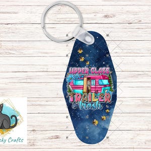 May include: A keychain with a navy blue background featuring a pink camper with the words "Upper Class Trailer Trash". The keychain has a silver ring and a white plastic connector. The design includes flowers, trees, and fireflies.