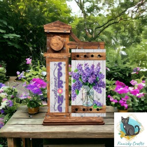 May include: Wooden welcome sign with the words "Welcome Spring" and a purple flower. The sign is attached to a wooden post with a decorative wooden panel featuring a bouquet of purple flowers in a glass jar.