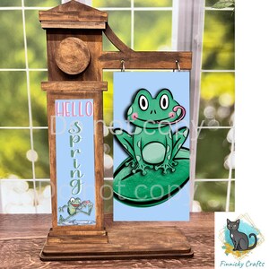 Puede incluir: Un letrero de madera con fondo azul y el texto "Hello Spring" en el lado izquierdo. En el lado derecho hay una rana verde con lengua rosa sentada en un nenúfar verde.