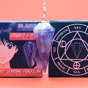 Pode incluir: Um pêndulo de adivinhação de ametista com uma corrente prateada pendura-se em frente a duas caixas. O pêndulo é roxo e transparente. Uma caixa apresenta arte de anime e o texto "AMETHYST DOWSING PENDULUM". A outra caixa tem um design geométrico com as palavras "YES" e "MAYBE".