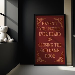 May include: A framed burgundy poster with gold lettering that reads "Haven't you people ever heard of closing the god damn door." A white teddy bear sits on a windowsill. The room is painted black.