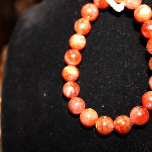 May include: A red sunstone beaded bracelet with a gold clasp. The beads are round and have a sparkly finish.
