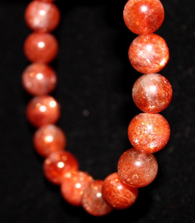 May include: A close-up of a sunstone bracelet with round, orange beads. The beads are polished and have a shimmery, iridescent finish.