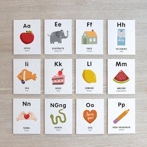 May include: Set of 12 flashcards featuring colorful illustrations and corresponding words in a specific language. Each card displays a letter of the alphabet with an image of an object. The cards include images of an apple, elephant, house, fish, cake, lemon, watermelon, hand, snake, love, and pencil.