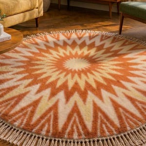 May include: A round area rug featuring a vibrant sunburst pattern in shades of burnt orange, cream, and pale yellow. The rug has a thick, textured pile and is finished with a decorative fringe border made of light beige tassels around the entire edge.
