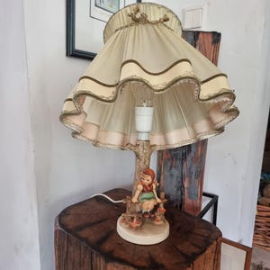 Może przedstawiać: Lampa stołowa z kremowym, plisowanym abażurem z tkaniny o falistych krawędziach i złotym wykończeniu. Podstawa to rzeźbiony pień drzewa z figurką osoby w czerwonym kapturze i zielonej sukience, siedzącej na drewnianym płocie.