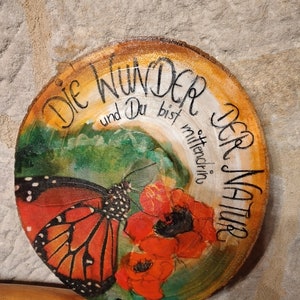 Puede incluir: Una rodaja de madera con una mariposa pintada, flores rojas y el texto "Die Wunder der Natur und Du bist mittendrin".