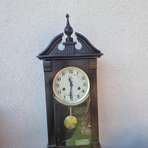 Puede incluir: Un reloj de pared de madera oscura con una esfera blanca y manecillas y péndulo dorados. El reloj tiene forma rectangular con una parte superior decorativa y una parte frontal de vidrio. La esfera muestra los números del 1 al 12.