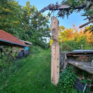 Könnte beinhalten: Eine hohe, helle Holzskulptur mit Löchern und farbigen Akzenten hängt an einem Holzbalken. Die Skulptur befindet sich in einer grünen Außenumgebung, mit einem Gebäude mit rotem Dach und Bäumen im Hintergrund. Die Skulptur ist etwa 1,8 Meter hoch.
