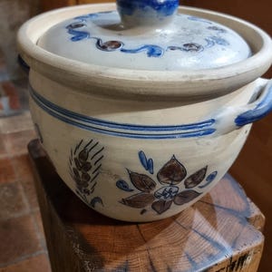 Könnte beinhalten: Ein weißer Keramiktopf mit blauem und braunem Blumendesign und einem blauen Knopfdeckel. Der Topf steht auf einem Holzstamm.