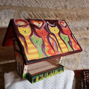 Può includere: Un mangiatoia per uccelli in legno dipinta con cinque gatti colorati con cuori sopra le loro teste. La mangiatoia ha un tetto rosso e un vassoio di legno per il mangime per uccelli.