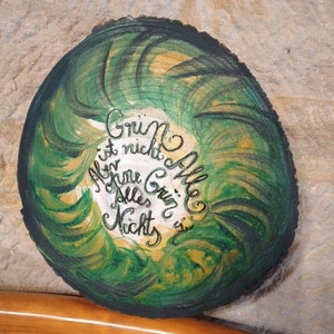 Puede incluir: Una placa redonda de madera con un diseño abstracto verde y dorado. La placa tiene una inscripción blanca que dice "Grin is not Hille, All the Grin is Alles Nicht".