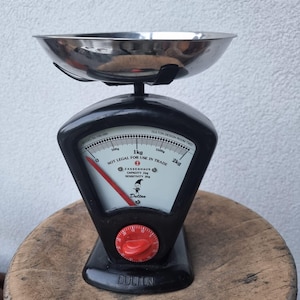 Op de afbeelding: Een vintage zwarte en zilveren keukenweegschaal met een roestvrijstalen kom. De weegschaal heeft een rode wijzerplaat en een witte wijzerplaat met zwarte markeringen, die tot 2 kg meet. De tekst "Dulton" is zichtbaar op de basis.